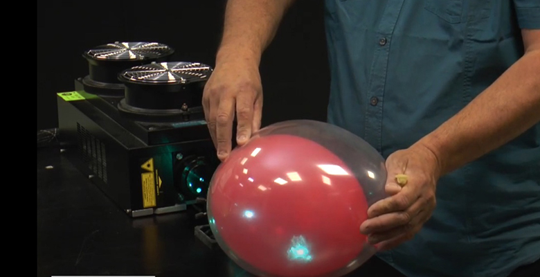 Visuel du SOT sur les lasers : chercheur en train de montrer le principe d'absoption en faisant éclater un ballon rouge à l'intérieur d'un ballon transparent par un laser bleu