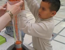 Photo montrant un petit garçon en train de remplir le tube en suspension au dessus d'une balance pour comprendre que le poids des grains est supporté par le tube et non par la balance.
